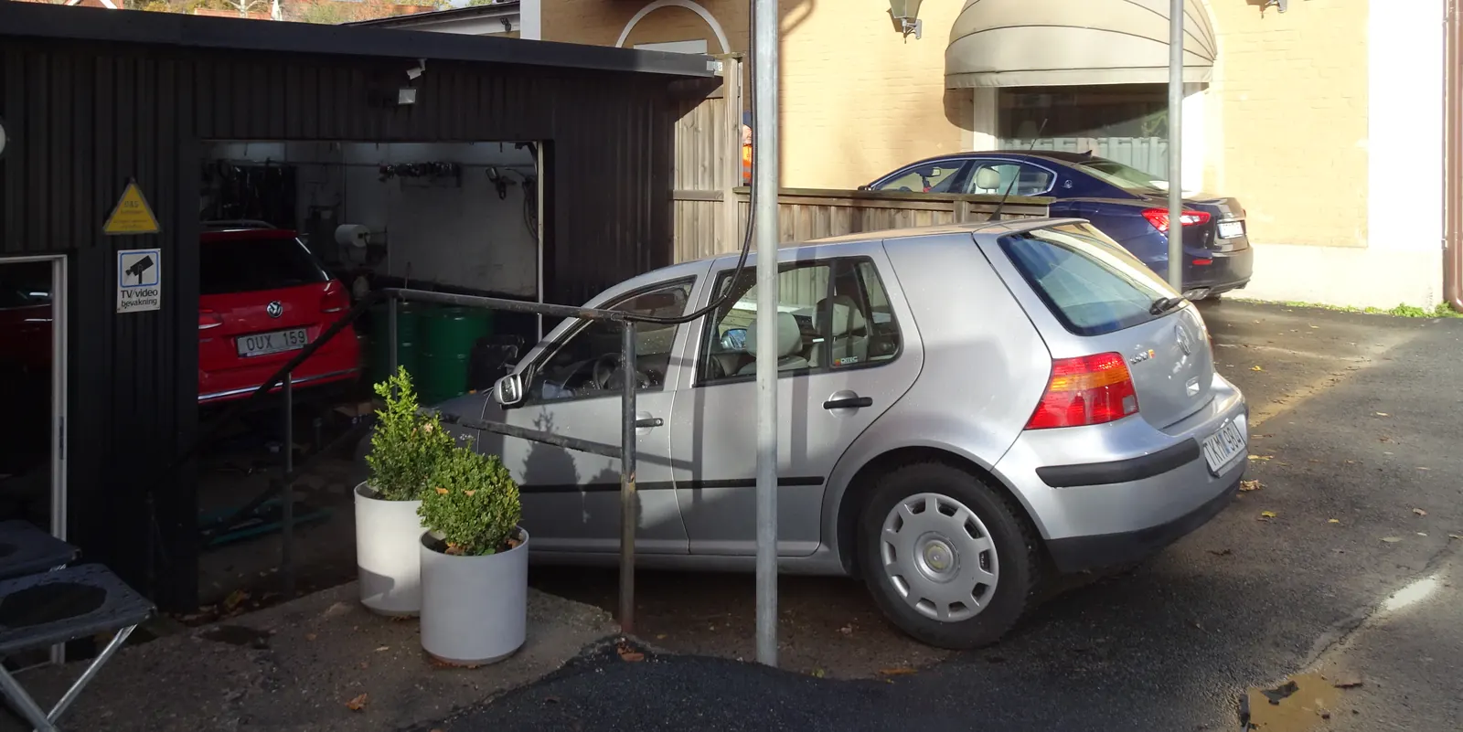 Öppen verkstadsport med en bil på lyft inne i verkstaden och bilar uppställda på gården utanför.